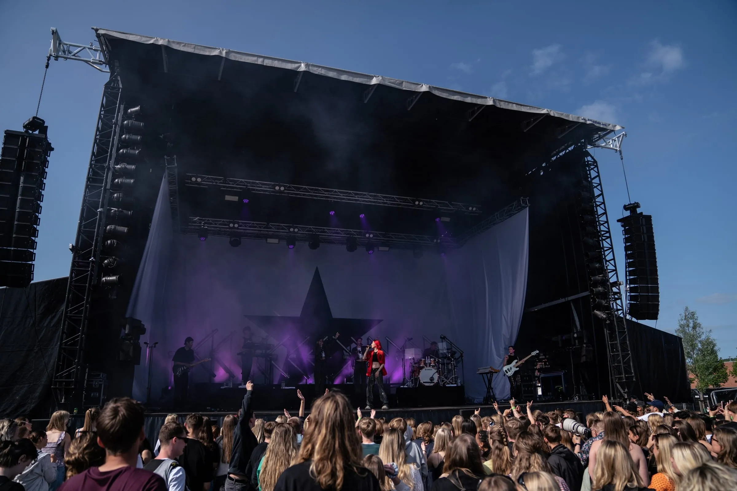 Scenen set skråt forfra i dagslys med publikum foran og røg i lyskeglerne