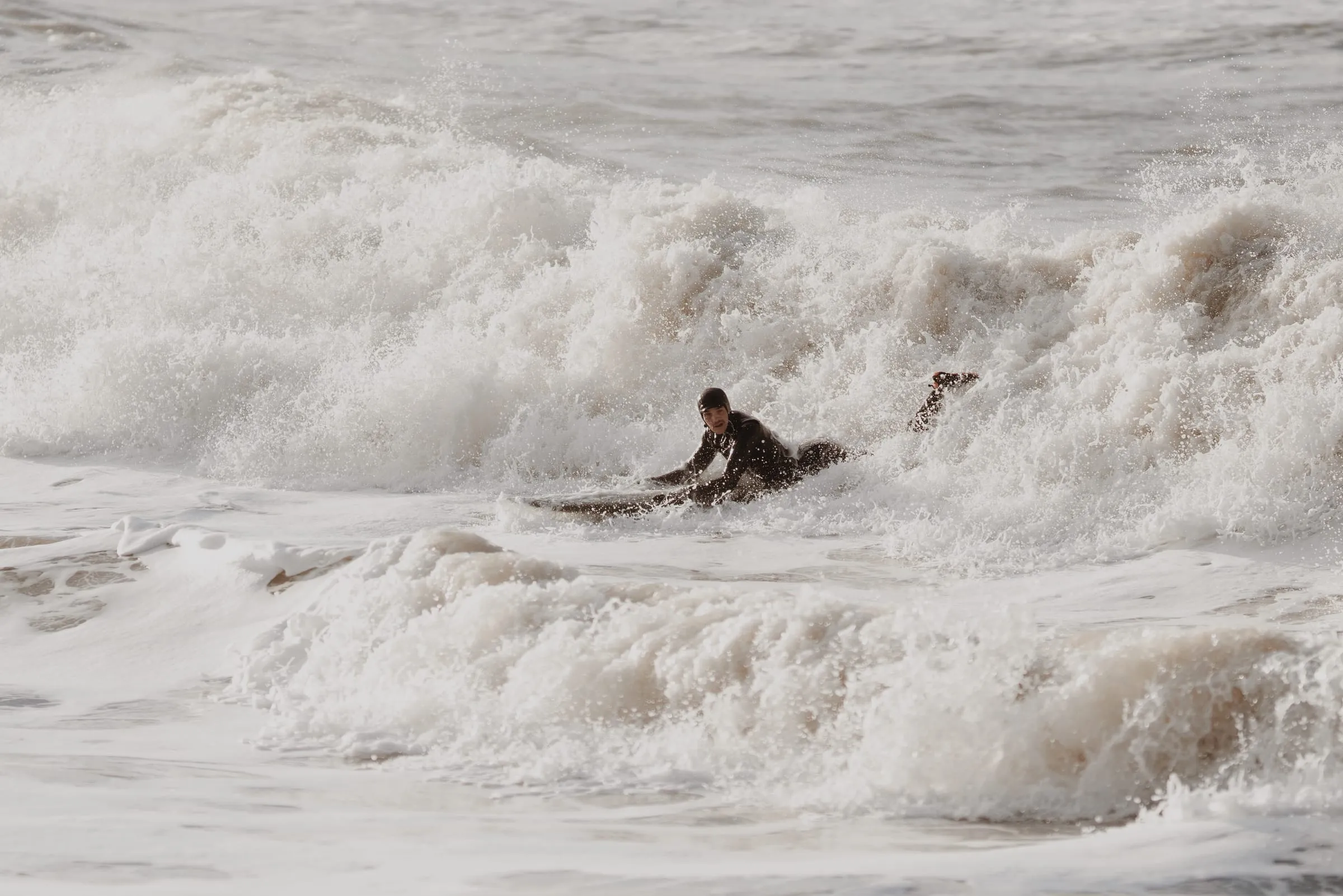 Surfer i sort våddragt kæmper sig ud gennem brydende skum i gråvejr