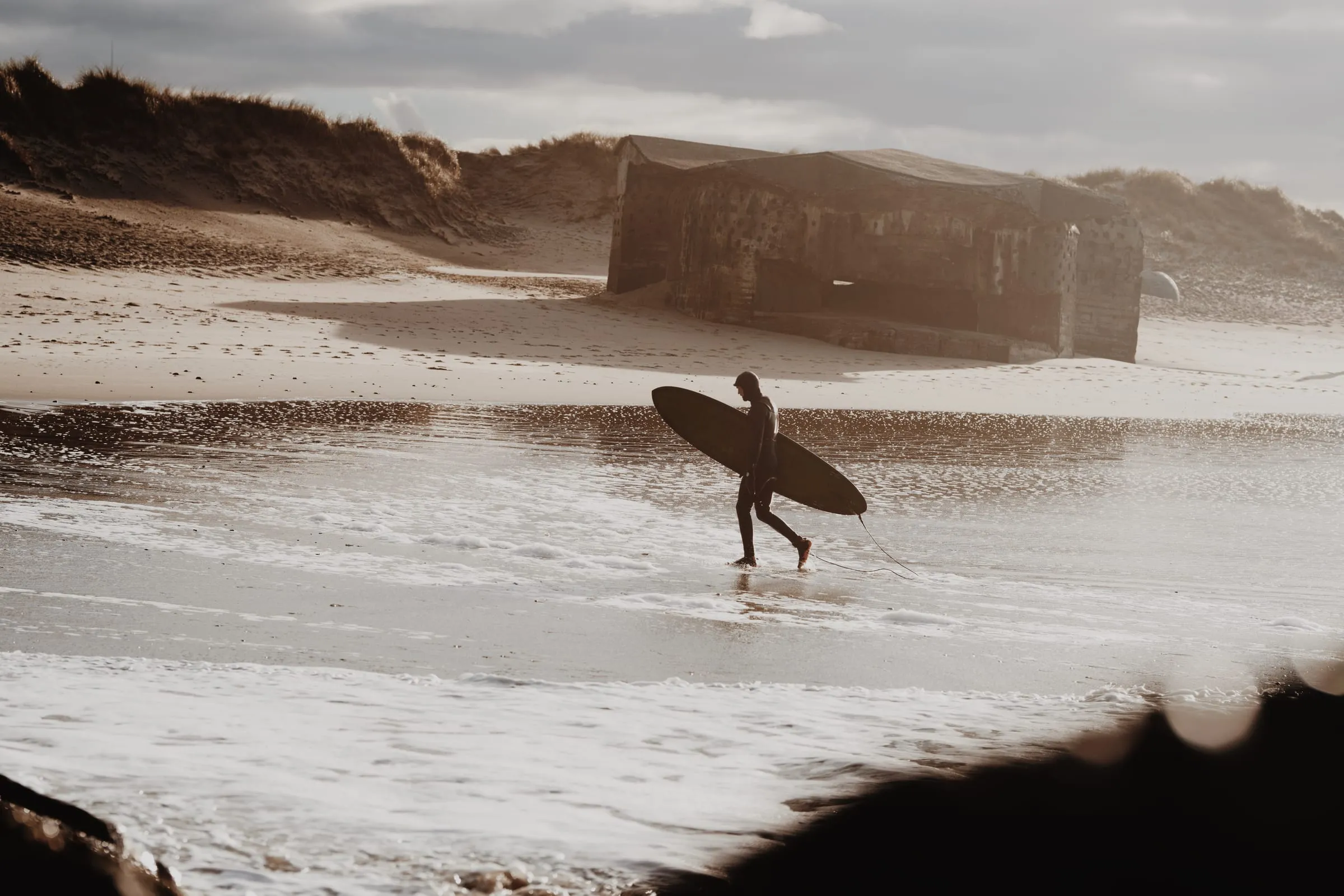 Surfer bærer brættet gennem lavvandet med bunkeren i klitten bagved