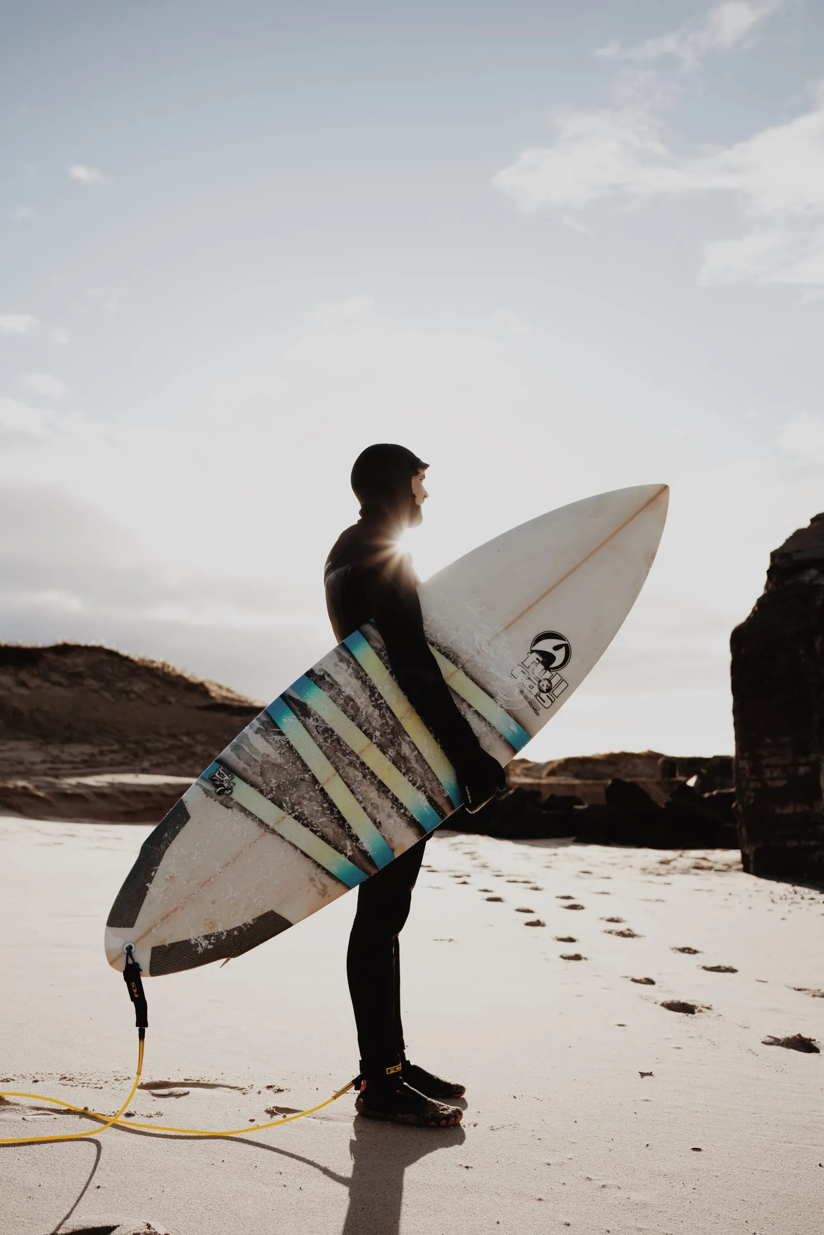 Surfer i hætte står med brættet under armen og den gule leash slæbende i sandet