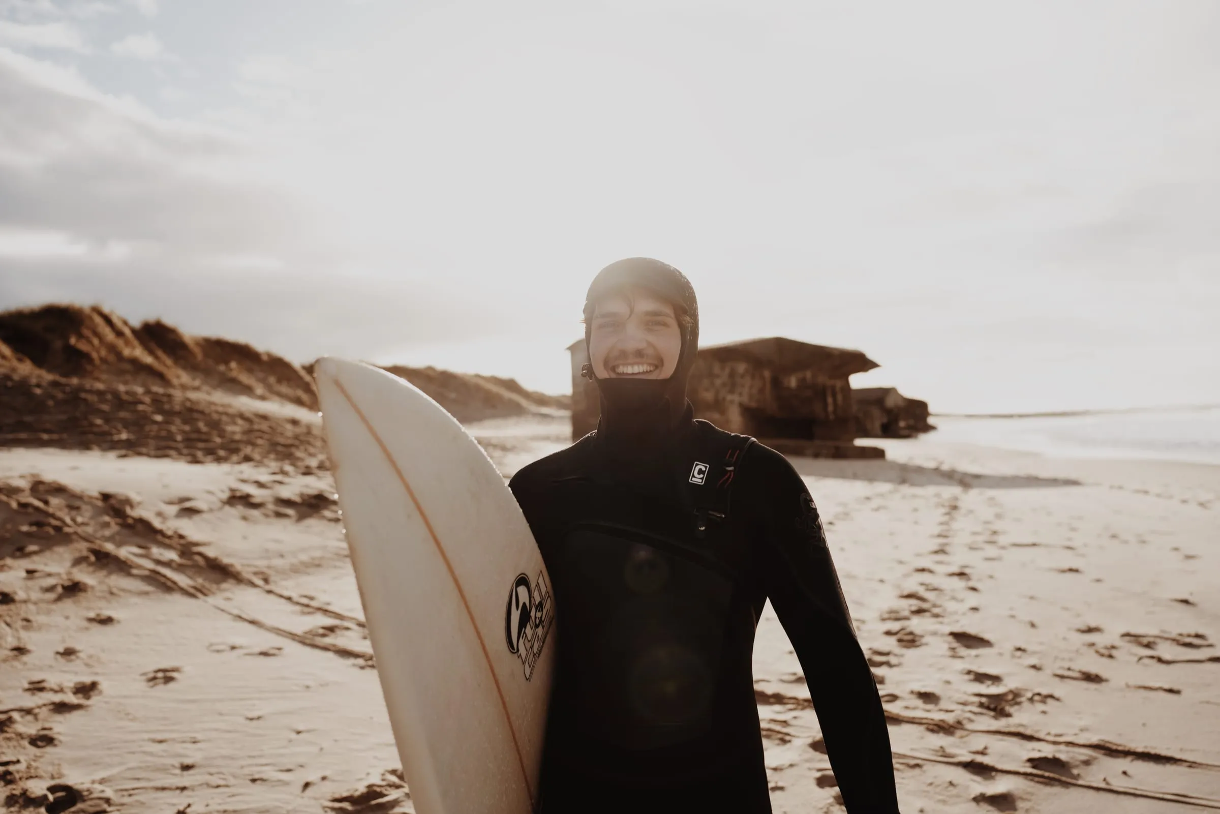 Surfer står med brættet på højkant og griner, bunkeren i baggrunden