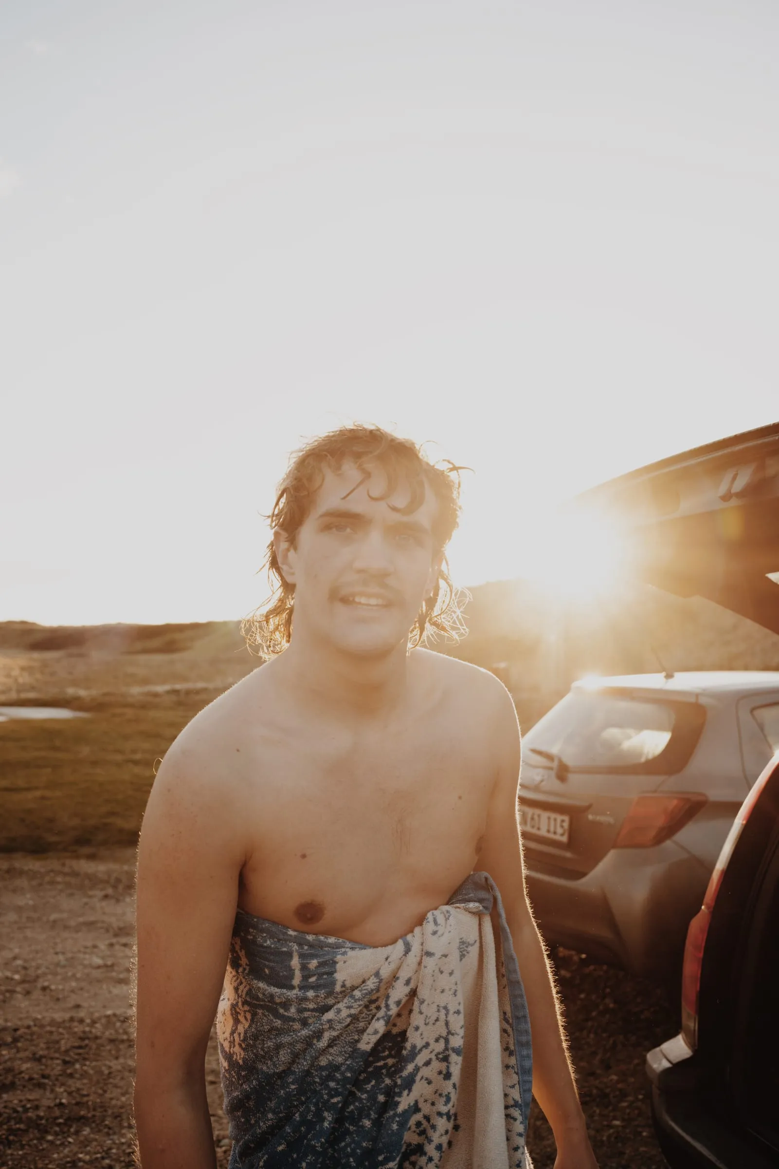 Surferen står med håndklædet om livet og solen nede bag bilen