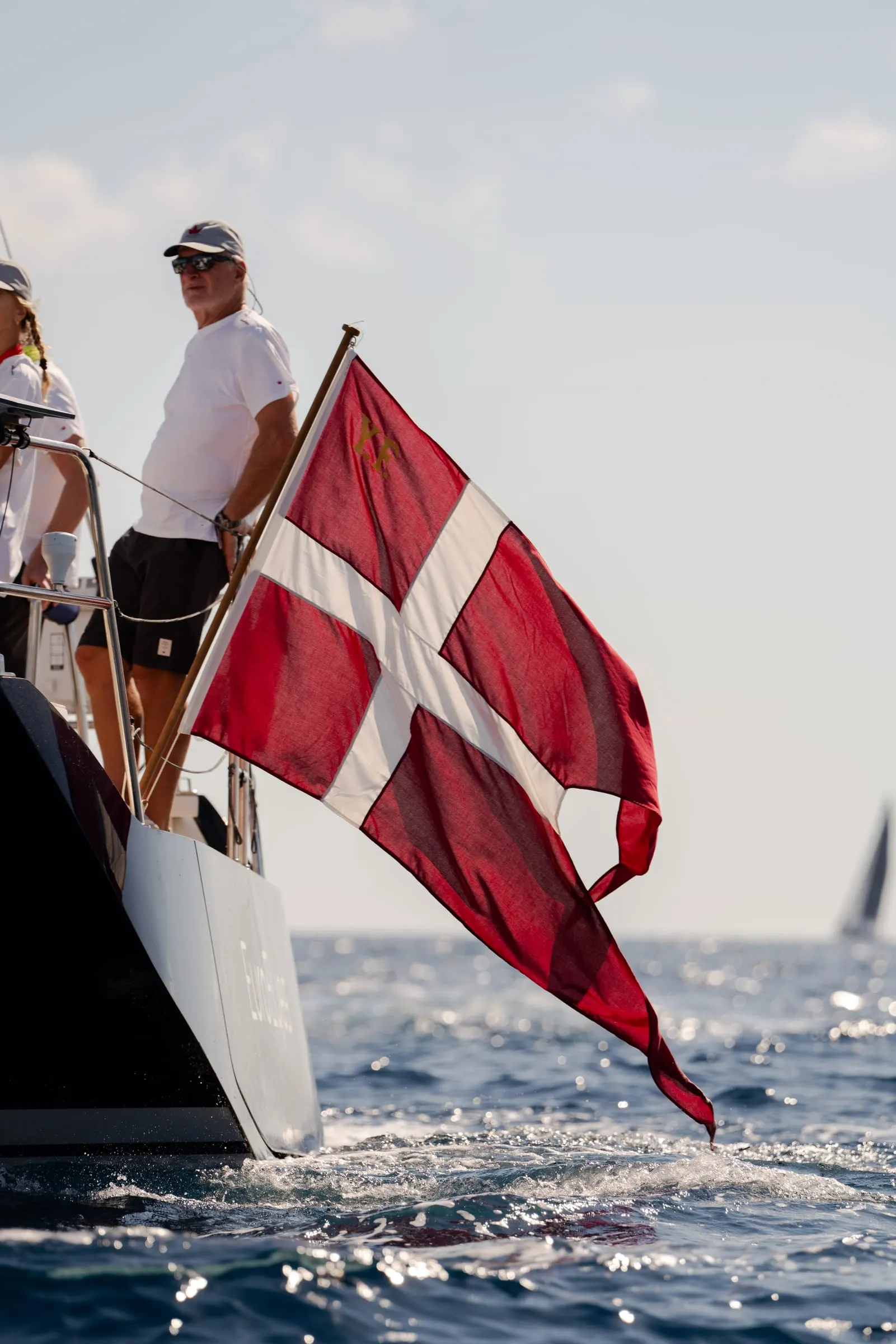 Dannebrog som yachtflag agter mens en gast i hvid T-shirt kigger ud over søen